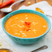 A bowl of creamy orange soup garnished with red bell pepper pieces, served on a wooden table.