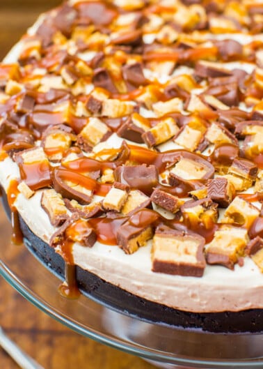 A chocolate cheesecake topped with whipped cream, caramel drizzle, and chopped chocolate bars.