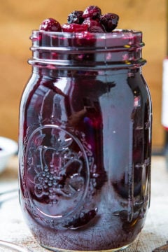 A glass jar filled with dark purple fruit jam, topped with whole berries and red wine cranberry sauce, on a wooden table.