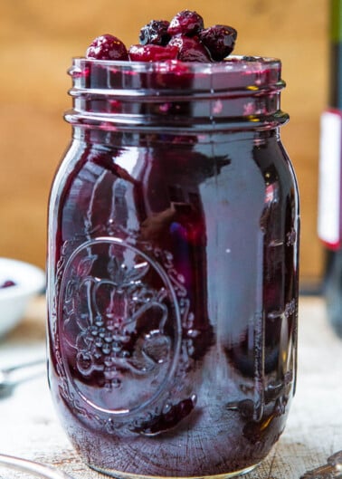 A glass jar filled with dark purple fruit jam, topped with whole berries and red wine cranberry sauce, on a wooden table.