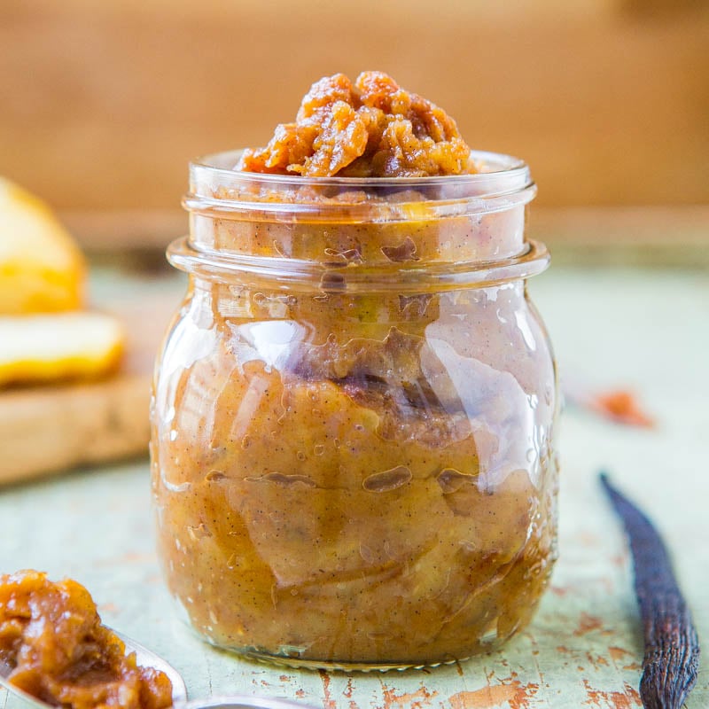 A jar of homemade apple butter on a rustic surface with slices of bread and a cinnamon stick.