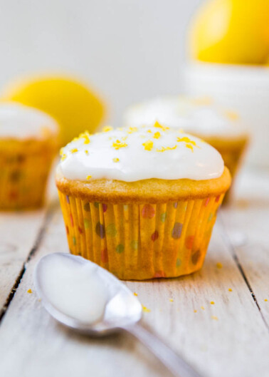 A lemon cupcake with white icing and zest garnish displayed on a wooden surface, with a spoon and more lemons in the background.
