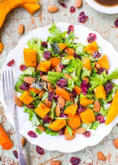 A colorful salad with butternut squash, mixed greens, cranberries, and almonds served on a white plate with a fork on the side.