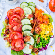 Colorful garden salad with tomatoes, cucumbers, carrots, corn, peas, lettuce, and bacon, served with a glass of milk.