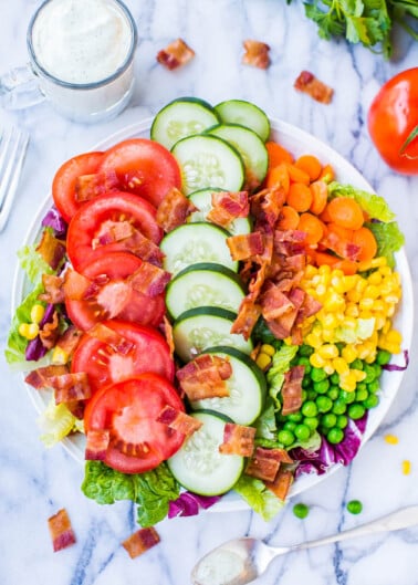 Colorful garden salad with tomatoes, cucumbers, carrots, corn, peas, lettuce, and bacon, served with a glass of milk.