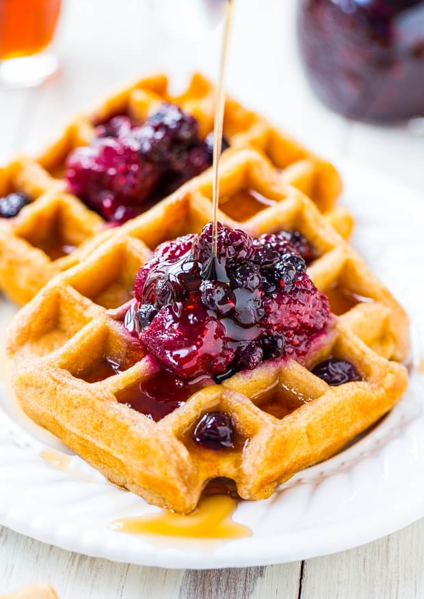 maple syrup being drizzled over two buttermilk waffles topped with berry preserves on a white plate