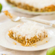 A slice of coconut pie on a white plate with the whole pie and green limes in the background.