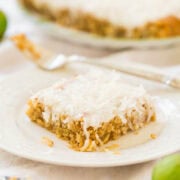 A slice of coconut pie on a white plate with the whole pie and green limes in the background.
