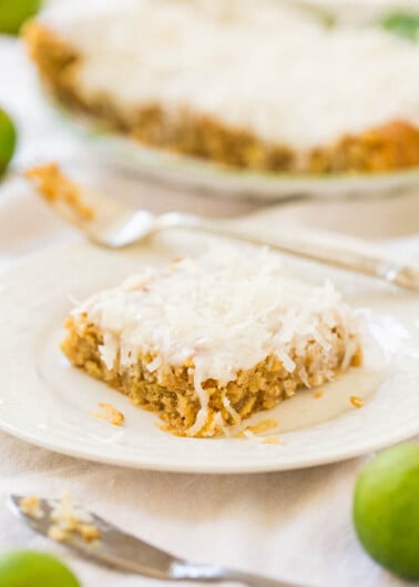 A slice of coconut pie on a white plate with the whole pie and green limes in the background.