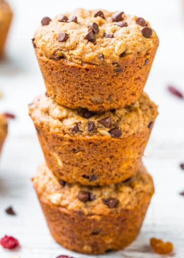 Three chocolate chip muffins stacked on top of each other on a white wooden surface.