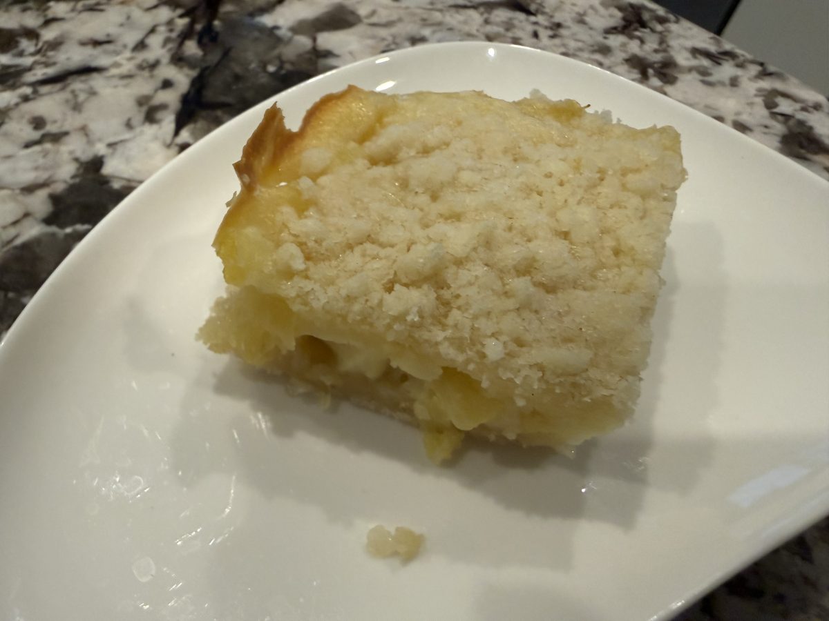 A square piece of crumb-topped cake with a creamy filling sits on a white plate on a marble countertop.