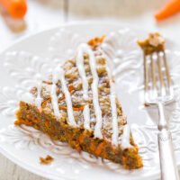 A slice of carrot cake with cream cheese frosting on an ornate white plate, accompanied by a fork.