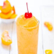 Peach smoothie topped with a cherry in a tall glass, with peach slices in the background.