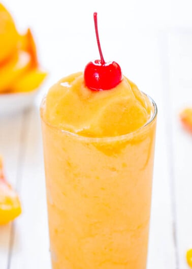 Peach smoothie topped with a cherry in a tall glass, with peach slices in the background.