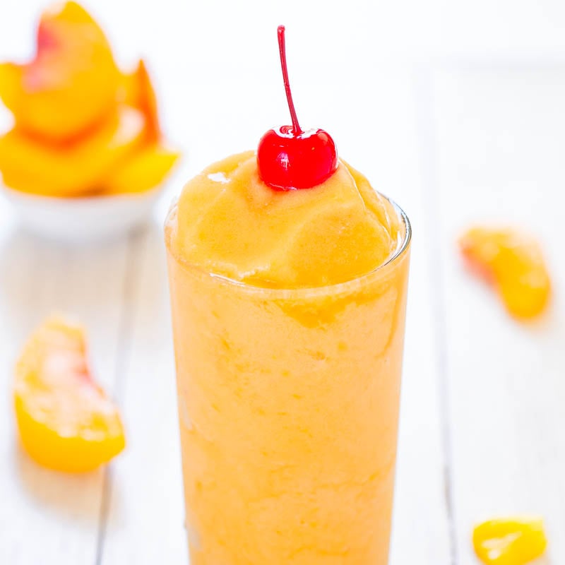 Peach smoothie topped with a cherry in a tall glass, with peach slices in the background.