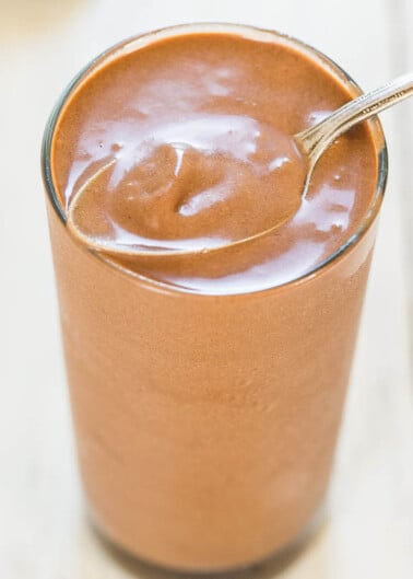 A glass of chocolate milkshake with a spoon on a wooden table.