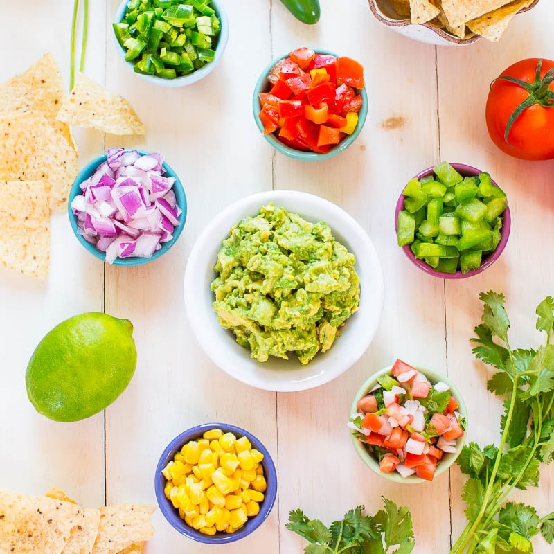 An assortment of fresh ingredients and a bowl of guacamole surrounded by tortilla chips on a wooden surface.