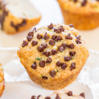 A close-up of a chocolate chip muffin on a white surface with more pastries blurred in the background.