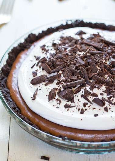 A chocolate pie topped with whipped cream and chocolate shavings, served on a clear glass dish.