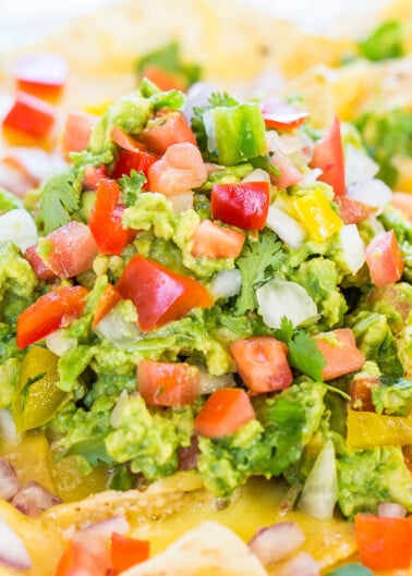 A close-up image of nachos topped with melted cheese, guacamole, diced tomatoes, onions, and cilantro.