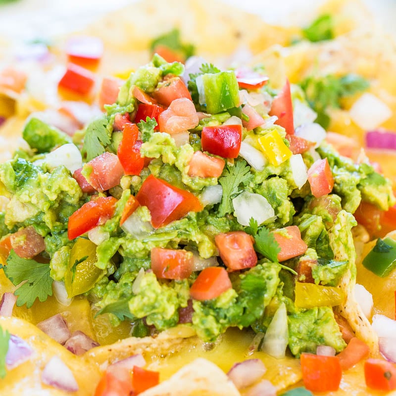 A close-up image of nachos topped with melted cheese, guacamole, diced tomatoes, onions, and cilantro.