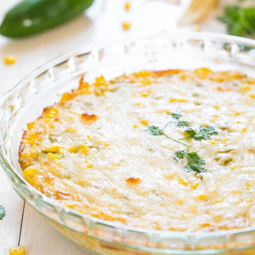 A round, clear glass dish containing a baked casserole topped with melted cheese and garnished with cilantro sits on a white surface, surrounded by scattered corn kernels and a green pepper in the background.