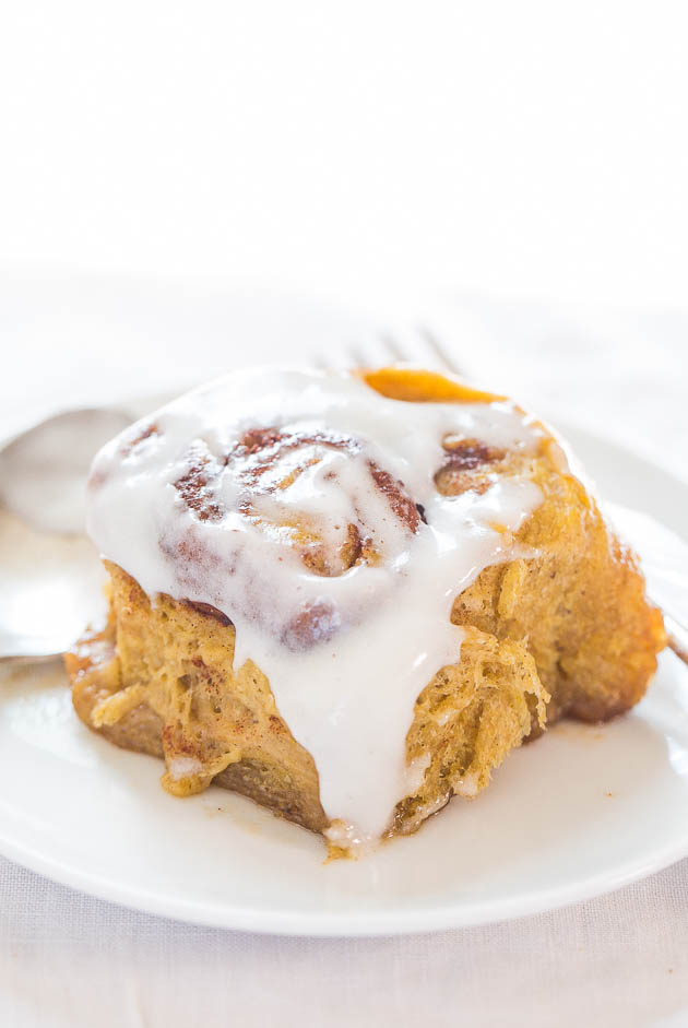 a glazed pumpkin cinnamon roll on a white plate. a fork rests in the background. 