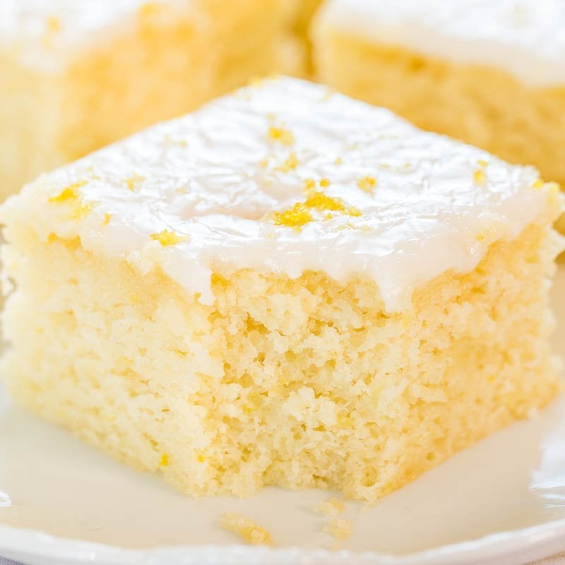 A close-up of a square piece of lemon cake with white icing and lemon zest on top.