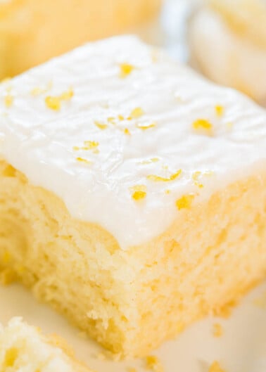 A close-up of a square piece of lemon cake with white icing and lemon zest on top.
