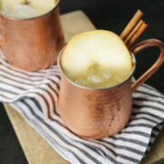 Two copper mugs with apple cider garnished with cinnamon sticks and apple slices on a wooden tray with a striped napkin.