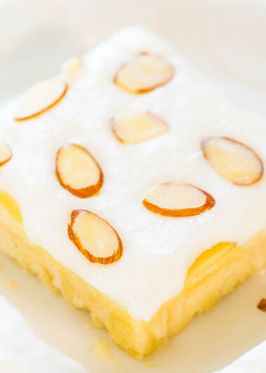 A close-up of a square piece of lemon cake topped with icing and almond slices on a white plate.