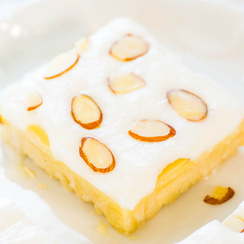 A close-up of a square piece of lemon cake topped with icing and almond slices on a white plate.