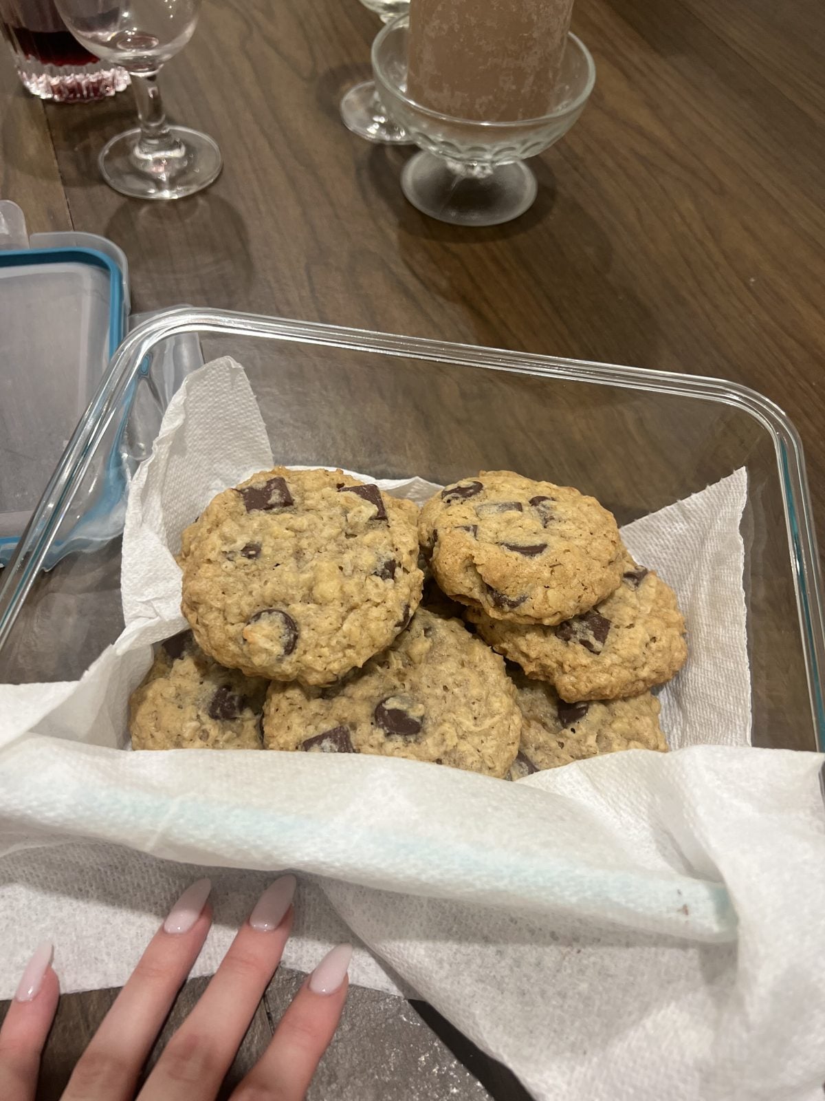 A glass container lined with paper towels holds six chocolate chip oatmeal cookies on a wooden table. A hand with manicured nails is touching the container.