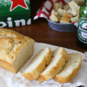 A loaf of bread sliced on a cutting board with a heineken beer bottle and a six-pack in the background.