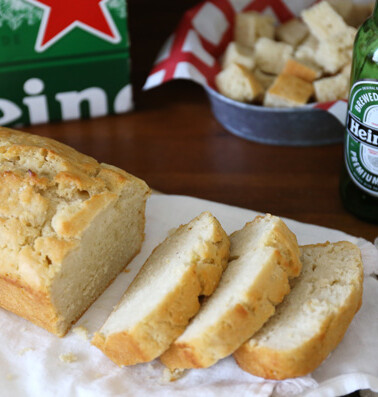 A loaf of bread sliced on a cutting board with a heineken beer bottle and a six-pack in the background.
