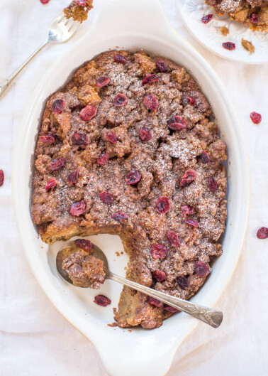 A baked bread pudding with cranberries dusted with powdered sugar in a white dish.