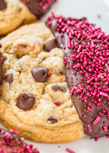 Chocolate chip cookies half-dipped in chocolate and adorned with pink sprinkles on a white plate.