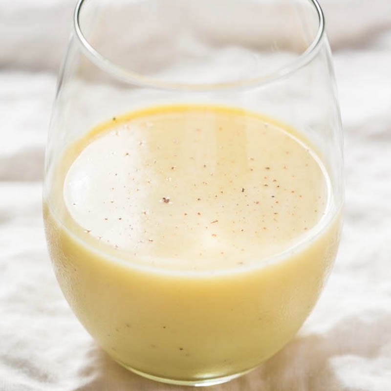 A glass of creamy smoothie with visible specks, possibly spices or seeds, on a light fabric surface.