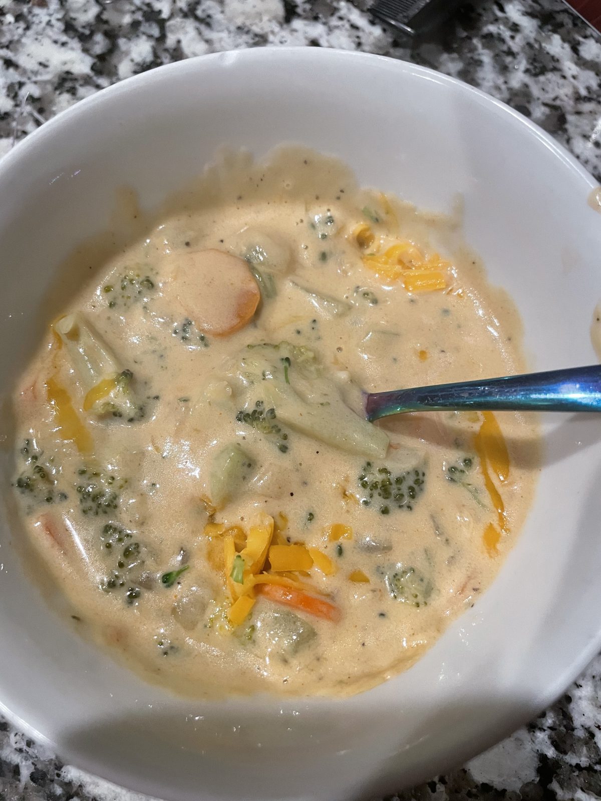 A bowl of creamy broccoli cheddar soup with visible pieces of broccoli, carrots, and cheddar cheese, on a granite countertop. A spoon is in the bowl.
