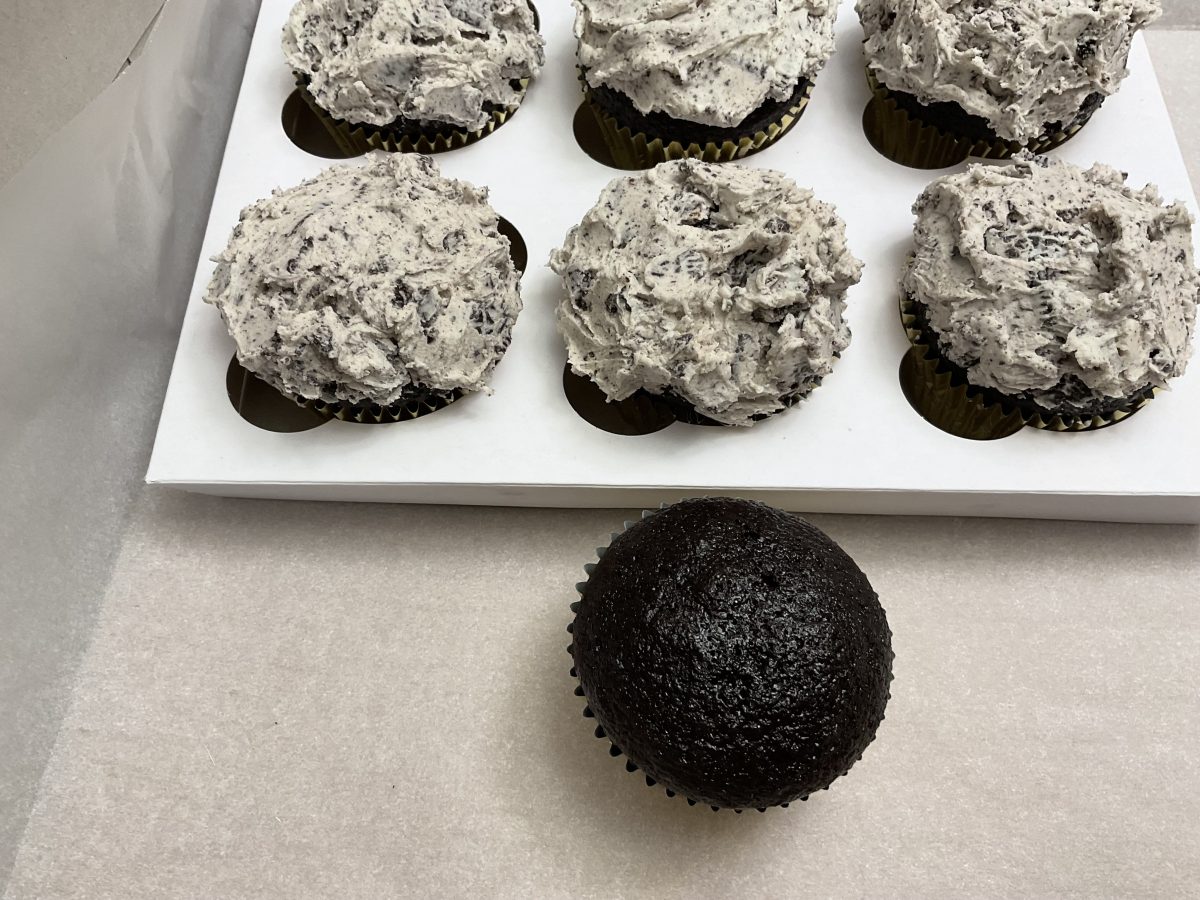 A single unfrosted chocolate cupcake sits on a surface in front of a box containing five chocolate cupcakes topped with cookies and cream frosting.