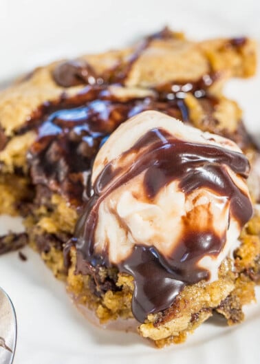 A scoop of vanilla ice cream melting over a warm chocolate chip cookie bar drizzled with chocolate syrup on a white plate with a spoon.