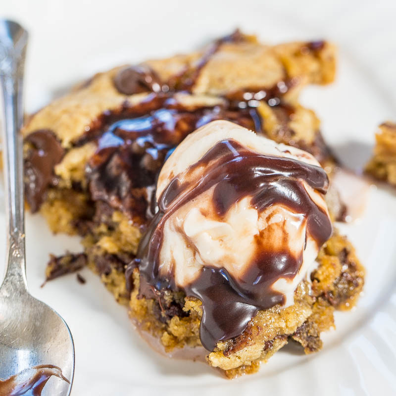 A scoop of vanilla ice cream melting over a warm chocolate chip cookie bar drizzled with chocolate syrup on a white plate with a spoon.