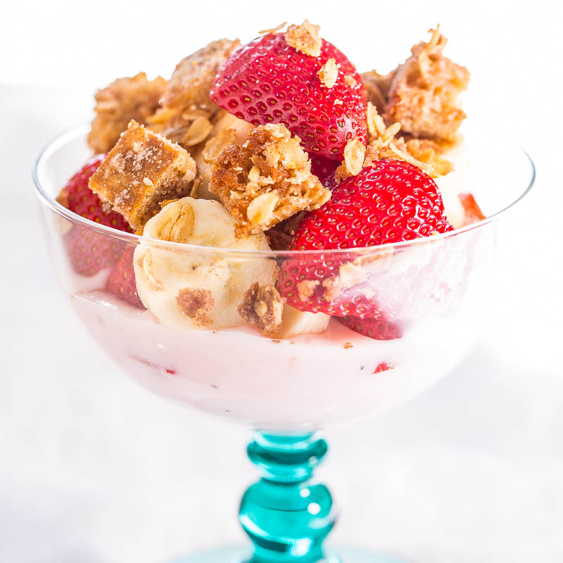 A glass dessert bowl filled with strawberry yogurt, fresh strawberries, banana slices, and topped with crunchy cereal pieces.
