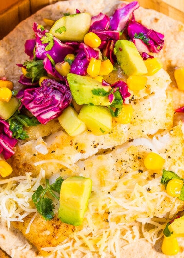 Colorful fish taco topped with avocado, purple cabbage, corn, and shredded cheese on a wooden surface.