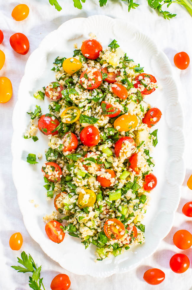 overhead view of tabbouleh on white platter surrounded by herbs and cherry tomatoes 