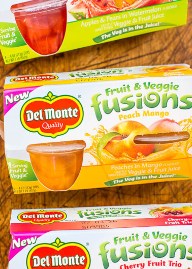 Packages of del monte fruit & veggie fusions snacks in peach mango and cherry fruit trio flavors displayed on a wooden surface.