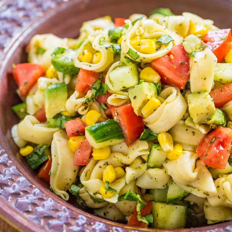 A bowl of pasta salad with tomatoes, cucumbers, avocado, and corn.