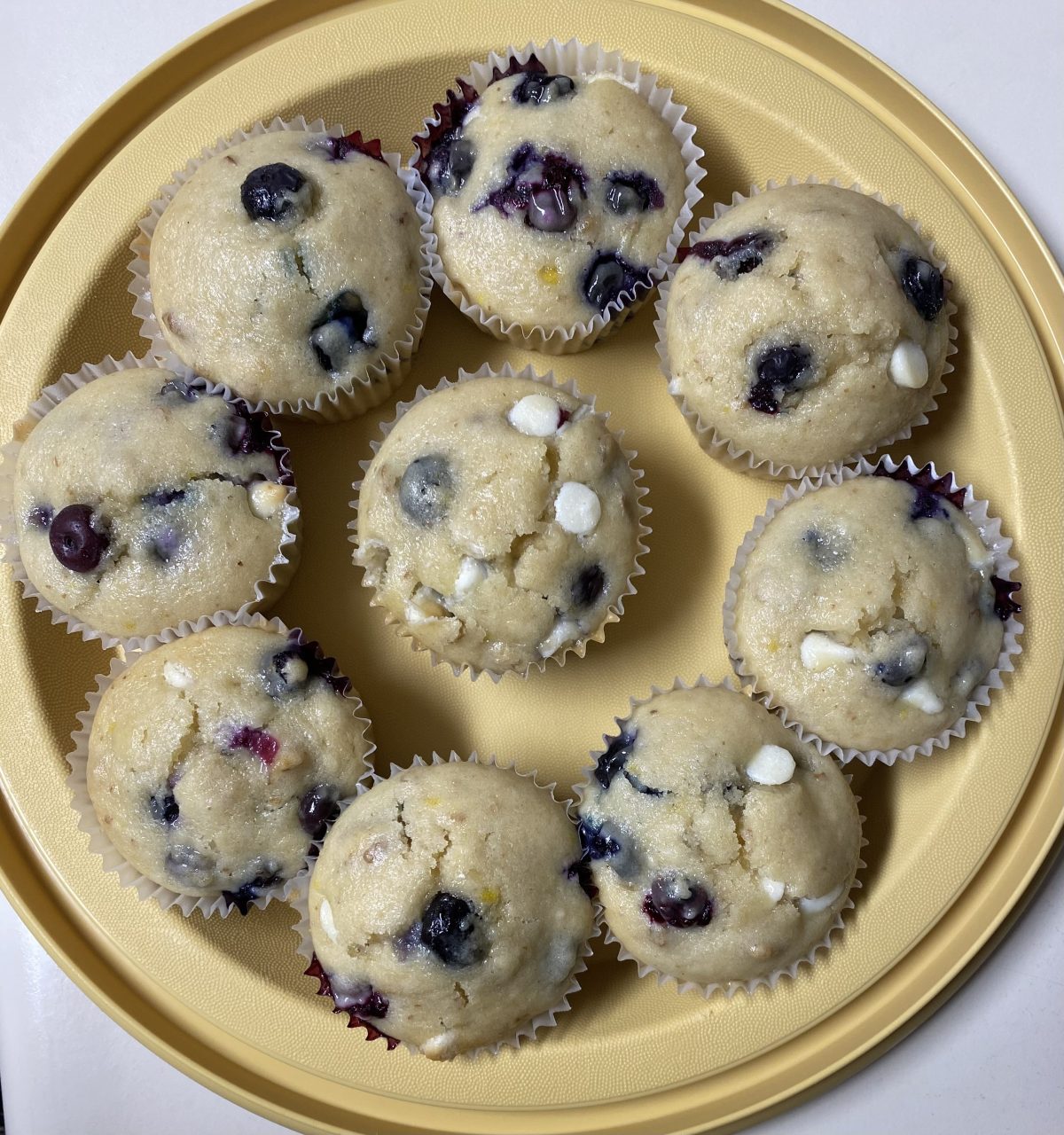 A round tray with ten blueberry muffins, some with white chocolate chips, arranged in a circular pattern on a white surface.