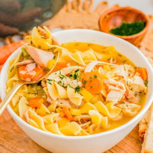 A bowl of chicken noodle soup with vegetables and herbs on a wooden board, accompanied by slices of bread and a small bowl of chopped parsley.