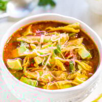 A bowl of chicken tortilla soup garnished with avocado slices and cheese, served on a wooden table.